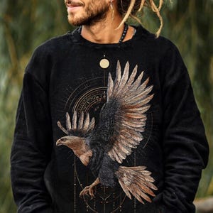 May include: A black long-sleeve sweatshirt featuring a large graphic of a soaring eagle with brown and black feathers. The design includes intricate geometric lines, circular patterns, and a small sun symbol positioned above the bird's wings.