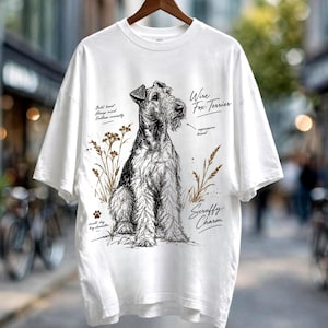 Könnte beinhalten: Ein weißes T-Shirt mit einer schwarz-weißen Skizze eines Wire Fox Terriers, umgeben von Weizenhalmen. Der Text lautet: Bold heart, Sharp mind, Endless curiosity, Wire Fox Terrier, expressive beard, Scruffy Charm und small dog big character.