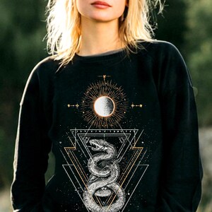 Op de afbeelding: Een zwart sweatshirt met lange mouwen en een grafische print van een opgerolde slang onder een zonicoon. Het ontwerp bevat geometrische driehoekige contouren, stralende lijnen en kleine steraccenten in witte en gouden inkt op de borst.