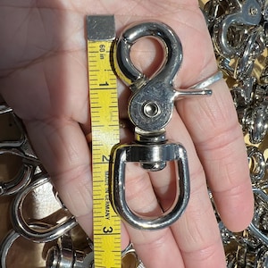 May include: A shiny, silver-colored swivel snap hook, approximately 3 inches long, with a spring-loaded gate. A yellow measuring tape is placed next to the hook. Other similar hooks are in the background.