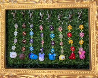 May include: A selection of beaded keychains with flower and character designs. Each keychain has a silver clasp, a beaded flower pattern in various colours, and a character charm. The charms include a white dog, a brown bear, and blue and purple bear-like characters.