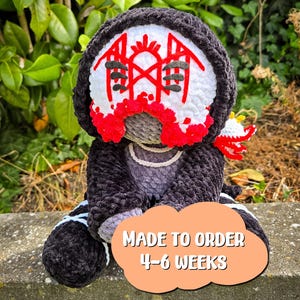 May include: A handmade crochet plush toy featuring a dark charcoal hooded cloak and a white face mask with intricate red and black embroidered symbols. The figure has a grey textured body, a white collar, and a small red and white accessory.