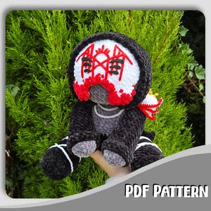 Op de afbeelding: Een gehaakte pluche pop met een zwarte capuchon, een wit gezichtsmasker met rood geometrisch borduurwerk en grijze ledematen. De pop heeft zwarte laarzen met witte kruissteken en een rood-wit accessoire op de rug. Tekst leest PDF PATTERN.