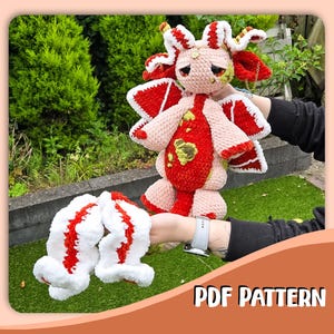 Op de afbeelding: Een gehaakt pluche wezen met een lichtroze kop, grote donkere ogen, een rood-wit patroon op het lichaam en grote vleugels. Het wezen heeft lange, golvende wit-rode aanhangsels en bevat de tekst PDF PATTERN aan de onderkant.