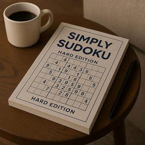 May include: A white mug filled with coffee sits next to a "Simply Sudoku Hard Edition" book and a black pen on a wooden table. The book features a grid with numbers, and the title is in blue.