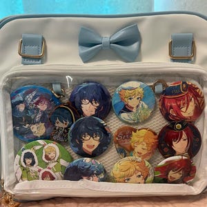 May include: White and light blue crossbody bag with a clear front pocket displaying several character buttons. The bag has a light blue bow and strap, with gold-colored hardware.