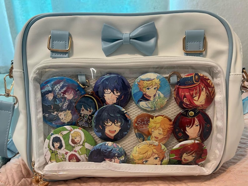 May include: White and light blue crossbody bag with a clear front pocket displaying several character buttons. The bag has a light blue bow and strap, with gold-colored hardware.
