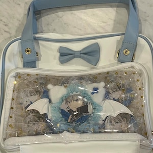 May include: White and light blue tote bag with a clear front pocket. The bag features a light blue bow and handles. The clear pocket displays character art, plushies, and a button. Gold accents are visible.