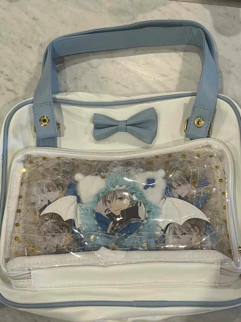 May include: White and light blue tote bag with a clear front pocket. The bag features a light blue bow and handles. The clear pocket displays character art, plushies, and a button. Gold accents are visible.