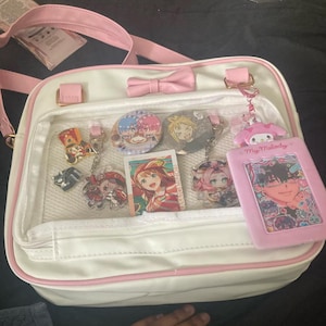 May include: White satchel bag with pink trim, a bow, and a clear front pocket displaying anime-style keychains, buttons, and a card. The bag has a pink strap and gold-colored hardware. A pink My Melody card holder is also visible.
