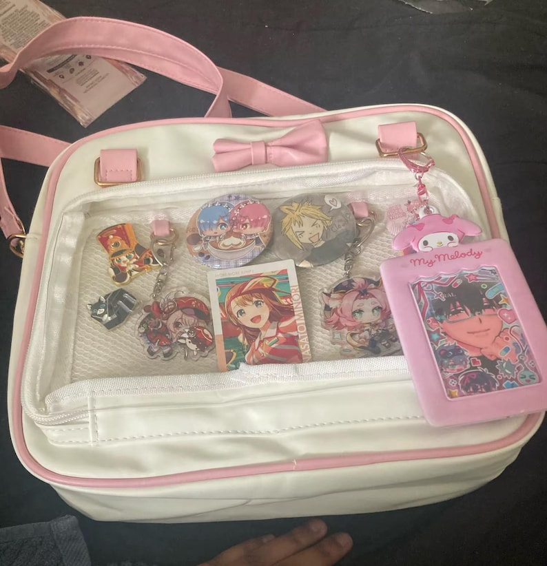 May include: White satchel bag with pink trim, a bow, and a clear front pocket displaying anime-style keychains, buttons, and a card. The bag has a pink strap and gold-colored hardware. A pink My Melody card holder is also visible.