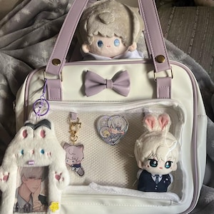 May include: A white tote bag with lavender accents, a bow, and a clear front pocket. The bag is filled with plush dolls and keychains. A plush doll with bunny ears and a plush doll with light-colored hair are visible.