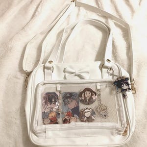 May include: White satchel bag with a bow detail and a clear front pocket displaying anime-style character pins and charms. The bag has a shoulder strap and a small keychain with a character charm.