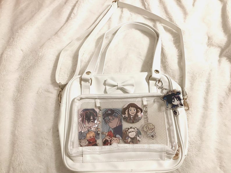 May include: White satchel bag with a bow detail and a clear front pocket displaying anime-style character pins and charms. The bag has a shoulder strap and a small keychain with a character charm.