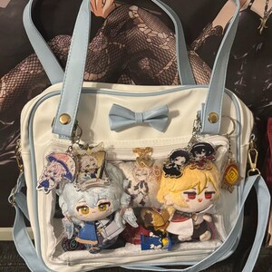 May include: White and light blue handbag with a bow and two handles. The bag has a clear front pocket with plush dolls and keychains. The bag has a light blue shoulder strap and a light blue bow.
