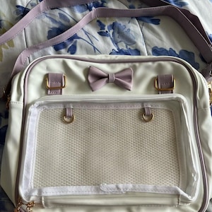 May include: White crossbody bag with a lavender bow and strap. The bag features gold-colored hardware and a clear, mesh-lined front pocket. The bag is rectangular and has a zipper closure. The bag is designed with a cute and feminine aesthetic.