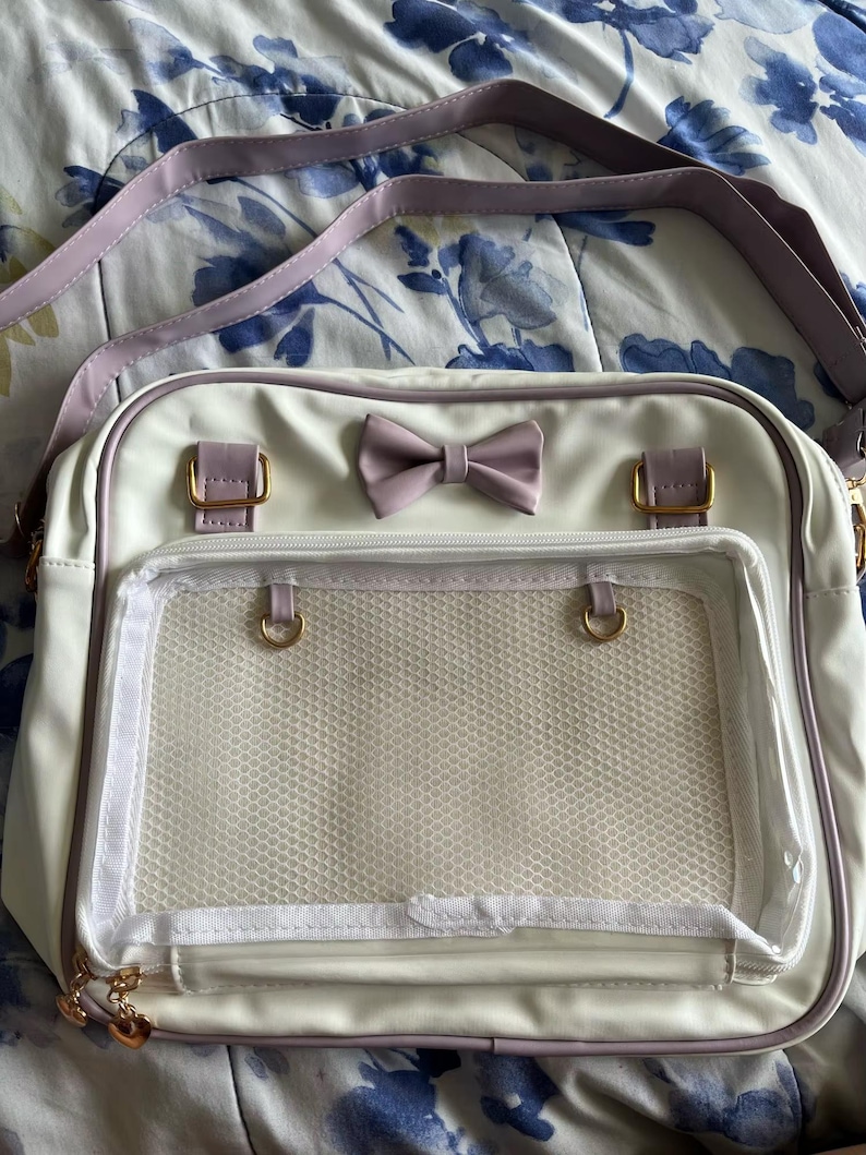 May include: White crossbody bag with a lavender bow and strap. The bag features gold-colored hardware and a clear, mesh-lined front pocket. The bag is rectangular and has a zipper closure. The bag is designed with a cute and feminine aesthetic.