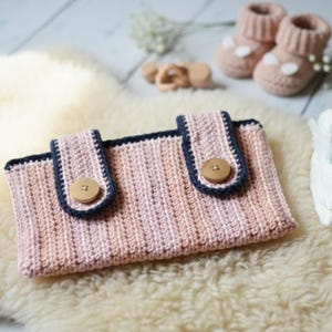 May include: A handmade, crochet baby clutch in shades of pink and peach, with navy blue trim and wooden button closures. The clutch is displayed on a fluffy, cream-colored surface, with matching baby booties and a wooden toy in the background.
