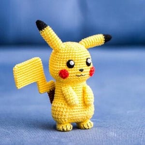 Puede incluir: Figura de Pikachu amarilla hecha a mano con ganchillo, con orejas con puntas negras, mejillas rojas y cola de rayo. La figura está sobre una superficie azul. El trabajo de ganchillo es detallado y es un personaje popular.