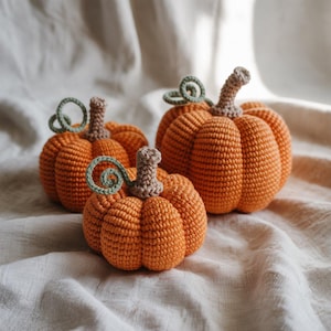 Peut inclure: Trois citrouilles crochetées de tailles différentes. Les citrouilles sont orange vif avec des surfaces texturées, des tiges marron et des vrilles vertes bouclées. Elles sont disposées sur un tissu de couleur neutre, créant une scène automnale et chaleureuse.