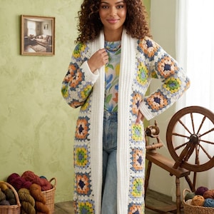 May include: A long, open-front cardigan with a white base and colourful square patterns. The squares feature orange, blue, yellow, and green accents. The cardigan is worn over a floral top and blue jeans. Baskets of yarn and a spinning wheel are in the background.