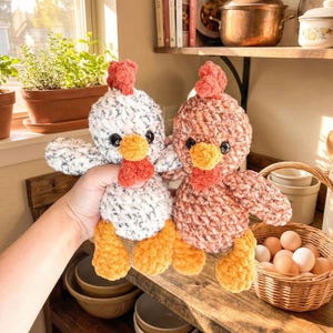 Puede incluir: Dos peluches de pollo hechos a mano con ganchillo. Uno es blanco con motas negras, el otro es de color marrón rojizo. Ambos tienen patas, picos y barbillas naranjas, y crestas rojas. Los pollos se exhiben en una estantería de madera cerca de una ventana.