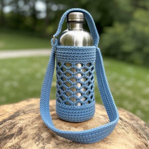 May include: A stainless steel water bottle in a light blue crochet carrier with a matching shoulder strap. The carrier has a lattice design, and the strap is adjustable. The bottle is resting on a wooden surface, with a blurred green background.