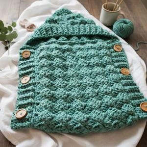 May include: A teal crocheted baby cocoon with a textured diamond pattern and a hood. The cocoon has wooden buttons along the side for closure. The item is displayed on a white cloth.