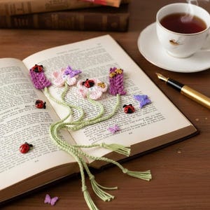 May include: An open book with a handmade crochet bookmark featuring purple, pink, and white flowers, ladybugs, and butterflies. A cup of tea, a pen, and other books are on the wooden surface.