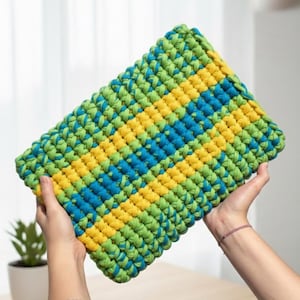 Może przedstawiać: Prostokątna, ręcznie szydełkowana kopertówka w żywym wzorze zieleni, żółci i błękitu. Gruba przędza tworzy teksturowaną powierzchnię. Kopertówka trzymana jest w dwóch dłoniach, prezentując ręczne rzemiosło.