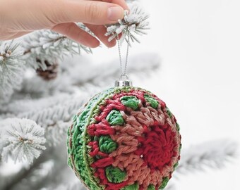May include: A handcrafted Christmas ornament with a crocheted design. The ornament features a combination of red, green, and pink yarn, creating a textured and festive appearance. It hangs from a silver string, ready to be displayed on a Christmas tree.