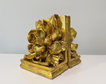 Puede incluir: Un par de sujetalibros de color dorado con un diseño de flor esculpida en tres dimensiones en la parte frontal. Los sujetalibros tienen un acabado metálico texturizado con reflejos y sombras visibles que resaltan los detalles de los pétalos.