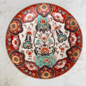 May include: A round decorative plate with a red border and intricate floral designs. The plate features a white center with green, orange, and black accents. Gold detailing adds a touch of elegance to this vintage-style piece.