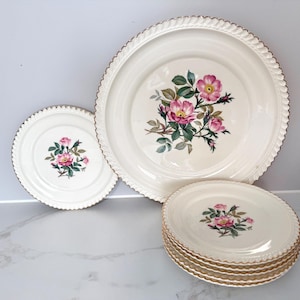 Pode incluir: Um conjunto de pratos vintage creme com um design floral de rosas. Os pratos têm uma borda dourada e uma borda ondulada. O conjunto inclui pratos grandes e pratos menores, perfeitos para um jantar formal.