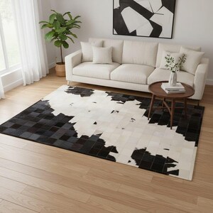 May include: A rectangular cowhide rug with a patchwork design. The rug features a mix of black, dark grey, and white squares, creating a geometric pattern. The rug is placed on a light-coloured hardwood floor.