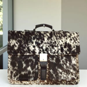May include: A briefcase with a spotted cowhide pattern in shades of black, brown, and white. The bag has a silver-colored clasp, a black leather strap, and a black leather handle. The briefcase is sitting on a white surface.