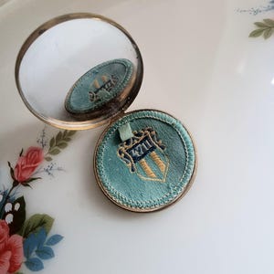 Może przedstawiać: Otwarty, zabytkowy kompaktowy lustro z okrągłą, złoconą ramką. Kompakt posiada wkładkę z jasnoniebieskiej tkaniny z haftowanym herbem w kolorze złotym i granatowym. Lustro odbija wnętrze kompaktu. Kompakt jest otwarty, ukazując lustro i pojemnik na puder.