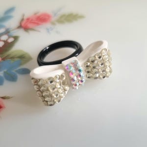 Könnte beinhalten: Ein weißer Schleifenring mit klaren Strasssteinen und schillernden Akzenten. Das Ringband ist schwarz. Die Schleife ist ein dekoratives Element, wahrscheinlich für Mode oder Schmuck.