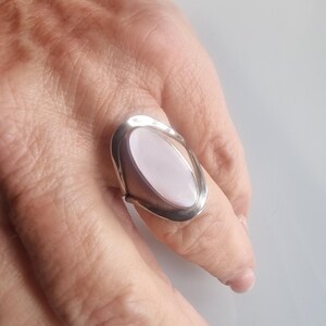 May include: A silver ring with an oval, light pink stone. The ring has a modern design with a polished silver band that curves around the stone. The ring is shown on a finger.