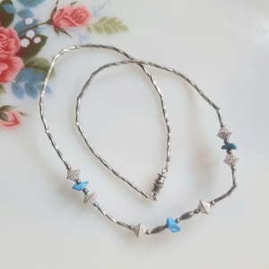 May include: A silver necklace featuring small, cylindrical beads and cone-shaped embellishments. The necklace is adorned with small, turquoise-coloured stones. The clasp is a simple barrel style. The necklace is displayed on a white surface.
