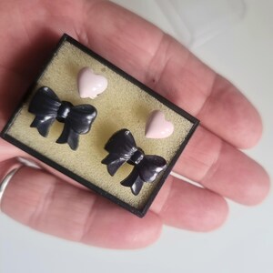 Könnte beinhalten: Ein Ohrring-Set in einer kleinen schwarzen Schachtel. Die Ohrringe umfassen zwei Paare: schwarze Schleifen-Ohrstecker und hellrosa herzförmige Ohrstecker. Die Schachtel hat eine gelbe Innenseite, und die Ohrringe werden darin präsentiert.