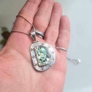 Puede incluir: Collar con colgante de plata con una concha de abulón iridiscente en el centro. El colgante tiene una forma irregular con grabados decorativos en los bordes. La cadena del collar es fina y plateada. Dimensiones: aprox. 3 cm.