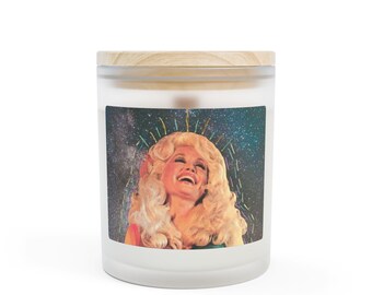 Puede incluir: Una vela blanca en un tarro de cristal con tapa de madera. La etiqueta presenta un collage de una mujer con cabello rubio riendo, sobre un fondo estrellado. La vela es un artículo de decoración para el hogar.