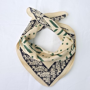 Op de afbeelding: Crèmekleurige bandana met een zwart en groen patroon. De bandana heeft een herhalend blad- en stippenpatroon, met een donkere rand. De stof is gevouwen en geknoopt, waarbij het ontwerp wordt getoond.