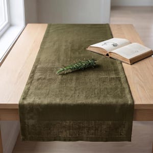 May include: A rectangular olive green table runner with a textured fabric finish and a darker border detail. An open hardcover book with cream-colored pages and a small sprig of green rosemary are placed on top of the runner on a wooden table.