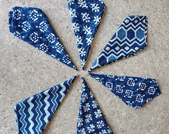 Puede incluir: Seis servilletas de tela azul índigo dispuestas en un patrón circular. Cada servilleta presenta un patrón blanco único, que incluye diseños florales, geométricos y punteados. Las servilletas están dobladas en forma triangular.