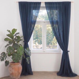 May include: A pair of floor-length, semi-sheer navy blue curtains hanging from a wooden rod over a window. The curtains are gathered and tied back on each side with matching fabric ties, creating a draped effect that allows light to filter through.