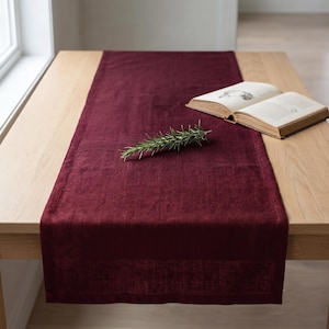 Può includere: Un runner da tavola rettangolare color bordeaux intenso con una superficie in tessuto testurizzato. Il runner è steso in piano e presenta un rametto di rosmarino verde al centro, accompagnato da un libro aperto con pagine color crema.