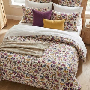 May include: A bed with a wooden frame is covered with a floral patterned duvet and pillowcases in shades of purple, yellow, and blue. Additional pillows in solid purple and yellow add color. A beige throw blanket is folded at the foot of the bed.