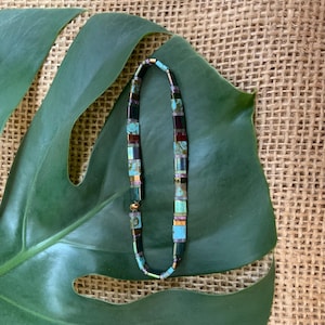 May include: A beaded necklace with turquoise, brown, and black rectangular beads. The necklace is displayed on a large green leaf against a burlap background. The beads are strung together with a gold-colored chain.